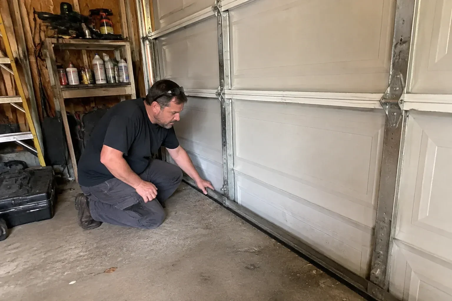 Person kneeling by a white paneled door in a garage, one hand on the black seal along its bottom edge. — Garage Door Roller Replacement, Brampton