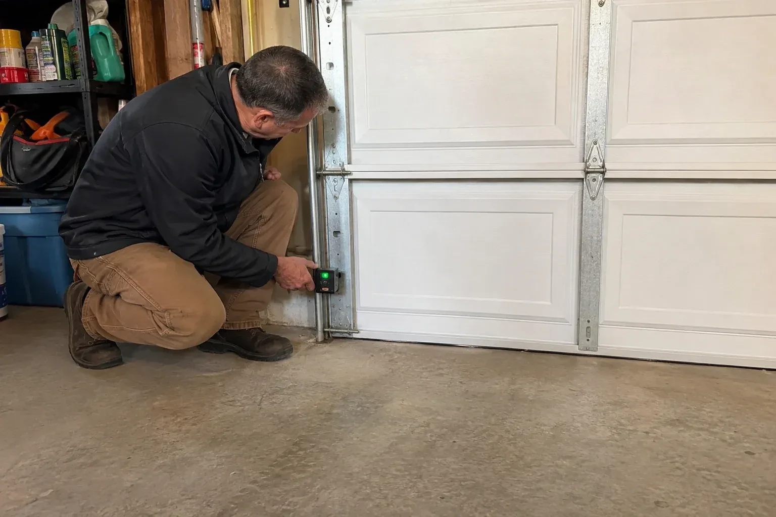 Technician kneeling beside a white paneled garage door, holding a small black box with a green light against the vertical track. — Photo-Eye Sensor Alignment or Replacement, Brampton