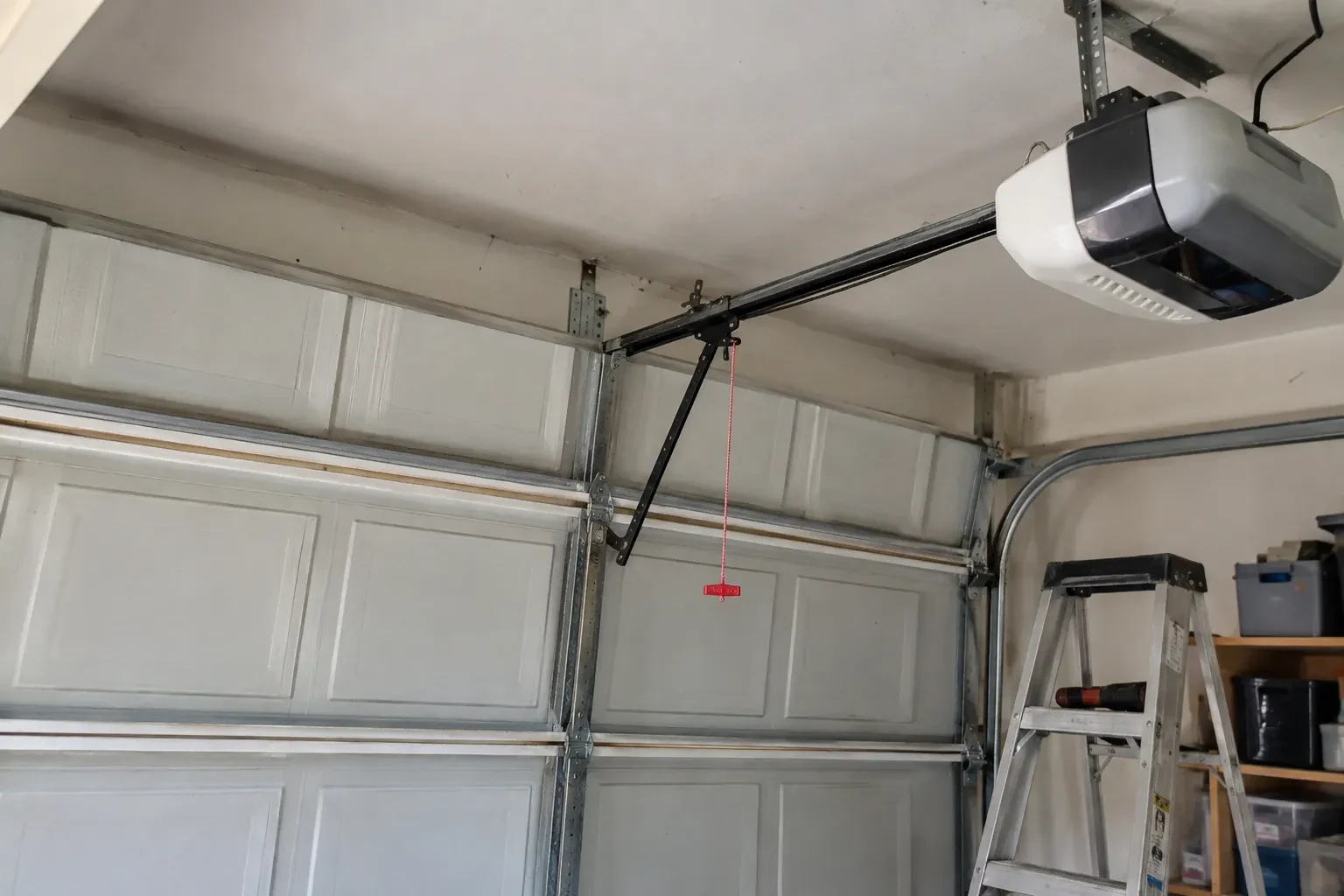 Garage-door opener on the ceiling with a bent black arm to a paneled door, a red release cord and a stepladder. — Brampton